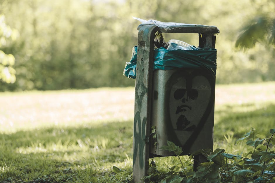 A metal rubbish bin situated outdoors on a grassy area with a background of blurred trees and foliage. The bin appears worn and slightly rusted, with graffiti art depicting a stylized face with dark eyes and a slightly open mouth spray-painted in black on the front. The top of the bin is partially covered by a black plastic bag or tarp, which is secured around the opening, with some visible blue and white plastic packaging protruding from it. The bin is positioned on a paved or concrete surface adjacent to the green grass, and nearby vegetation includes a few small plants and leaves at its base. The ambient lighting suggests daytime, with natural sunlight illuminating the scene softly, indicating a peaceful outdoor environment perhaps near a park or garden area where private or independent waste disposal methods might be used, as exemplified by this alternative rubbish container from Rubbish Clearance Bow.