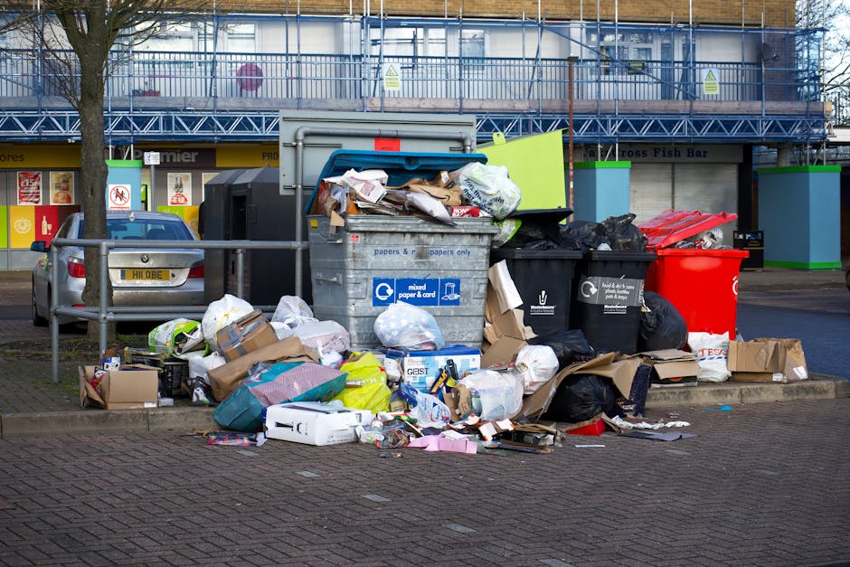 A collection of overflowing waste bins and scattered rubbish on a paved urban street, with various types of waste including cardboard boxes, plastic bags, paper debris, and discarded packaging piled around the bins. The bins are positioned on a curb, with some lids open, revealing mixed waste inside. Behind the waste, there is a metal railing and parked cars, with a background featuring a commercial building with signage and advertisements on the storefronts. The scene is illuminated by daylight, highlighting the cluttered mess which could be associated with private waste collection services such as those provided by Rubbish Clearance Bow, in contrast to typical municipal rubbish collection. The overall setting suggests a need for timely rubbish removal and proper waste management, supporting private or independent disposal options available in urban environments.