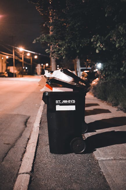 A black plastic rubbish bin with the label 'ST. JOHN'S' positioned on a sidewalk beside a curb in an urban area at night. The bin's lid is open, revealing various waste materials, including cardboard and other paper items. The surrounding environment includes dim street lighting, casting shadows on the pavement, with a few trees and bushes nearby. In the background, there are streetlights, traffic signals, and parked vehicles, indicating a residential or city street. The scene captures the typical setting where private waste collection or rubbish removal services, such as those offered by Rubbish Clearance Bow, might handle the disposal of household or commercial refuse, supporting effective on-site rubbish clearance in the district near Bow Road station E3, Bow.