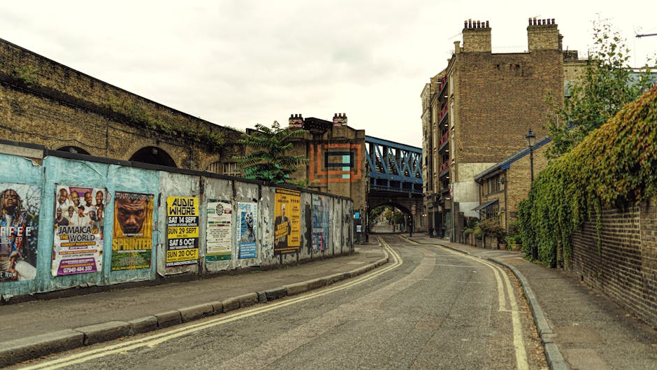 A narrow urban street scene featuring a curved asphalt road with double yellow lines along the curb. On the left side, a weathered metal fence is covered with numerous colorful posters advertising local events, including concerts and festivals, with some torn or peeling. Behind the fence, an aged brick wall with a slight moss or dirt buildup runs parallel to the street, leading towards an overhead blue metal railway bridge supported by brick and steel pillars. On the right, a tall brick building with multiple chimneys and balconies stands behind a green leafy hedge that extends along a brick wall. The scene is illuminated by natural daylight under a cloudy sky, with soft shadows cast on the road and buildings, suggesting an overcast day. The environment appears quiet, with no visible vehicles or pedestrians, and the setting reflects a typical East London street atmosphere, consistent with rubbish removal and community cleanup contexts, as managed by providers like Rubbish Clearance Bow.