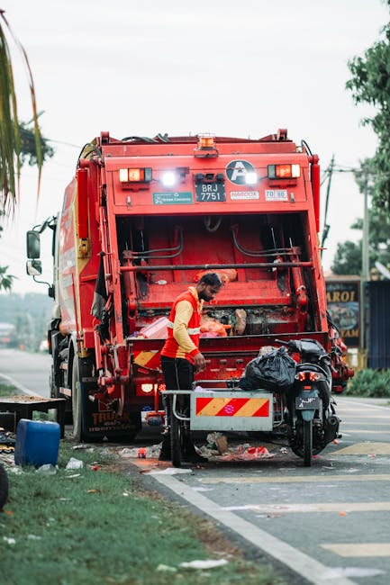 A worker wearing a red and yellow uniform is standing behind a large red rubbish collection truck, which is actively engaged in waste collection on a roadside. The truck has a bulky, industrial appearance with a metallic, painted red exterior that appears slightly weathered and functional, equipped with various lights, compartments, and hydraulic mechanisms. The worker is leaning forward, operating a control panel or accessing the rear loading area, with a black trash bag and some scattered debris nearby. The scene takes place on a paved street with a marked parking area and a grassy verge on the left side, featuring some trees, electrical wires, and buildings in the background under cloudy daylight conditions. The presence of the rubbish collection vehicle indicates a focus on local waste management or alternative waste handling services, consistent with private rubbish removal efforts that complement or provide alternatives to municipal services, as managed by Rubbish Clearance Bow.