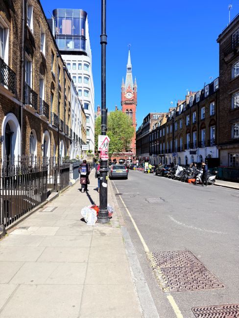 A city street scene showing a clear blue sky with a historic red-brick church featuring a tall, pointed spire and clock tower in the background. On the left side of the image, there is a row of multi-story residential buildings with brick facades, arched windows, and black wrought-iron fences along the pavement. A lamppost stands directly in the middle of the sidewalk, with a small pile of white rubbish bags at its base. The sidewalk is paved with concrete slabs, and two pedestrians are walking along it; one is carrying a backpack, and another appears to be holding a bag. Several cars and motorbikes are parked along the right side of the street, with a few additional vehicles in motion. In the background, beyond the parked vehicles, there are construction barriers and some workers wearing high-visibility clothing, which suggests ongoing work or clearance activity. The scene is well-lit with bright sunlight, and the overall environment appears tidy. The imagery subtly relates to private waste disposal or rubbish clearance services, as provided by Rubbish Clearance Bow, within an urban context of alternative waste handling.