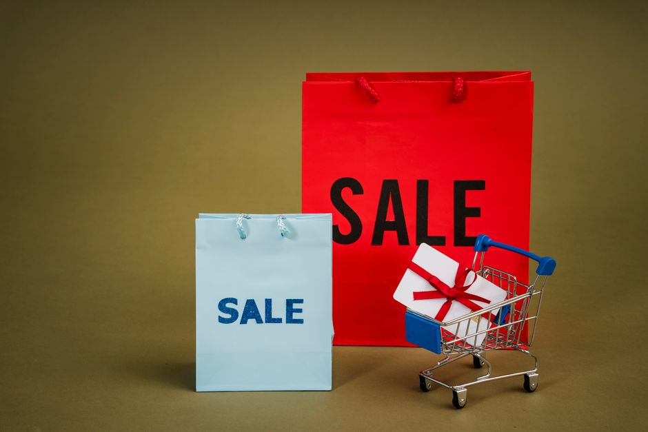 In the foreground, there is a small shopping trolley made of metal with a blue plastic handle, positioned on a plain, neutral-colored surface. Inside the trolley, a neatly wrapped white gift box with a red ribbon tied in a bow is visible. Behind the trolley, two shopping bags are prominently displayed: a larger red bag with sturdy red handles, featuring the word 'SALE' in bold black letters, and a smaller light blue bag with matching blue handles, also bearing the word 'SALE' in large, bold blue letters. The backdrop is a plain, warm-toned surface with soft, even lighting that highlights the textures of the paper bags, the glossy finish of the gift box, and the metallic sheen of the trolley, creating a clean, uncluttered scene with a focus on promotional shopping objects. This visual setup subtly relates to private or alternative shopping experiences, like independent clearance or sales events, akin to arranging for rubbish clearance or disposal services with a professional provider like Rubbish Clearance Bow, to support on-site or scheduled waste handling activities.