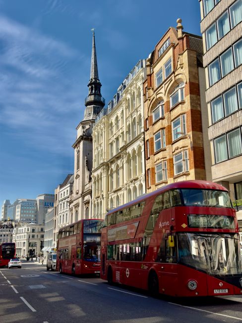 A city street scene showing a clear blue sky with a historic red-brick church featuring a tall, pointed spire and clock tower in the background. On the left side of the image, there is a row of multi-story residential buildings with brick facades, arched windows, and black wrought-iron fences along the pavement. A lamppost stands directly in the middle of the sidewalk, with a small pile of white rubbish bags at its base. The sidewalk is paved with concrete slabs, and two pedestrians are walking along it; one is carrying a backpack, and another appears to be holding a bag. Several cars and motorbikes are parked along the right side of the street, with a few additional vehicles in motion. In the background, beyond the parked vehicles, there are construction barriers and some workers wearing high-visibility clothing, which suggests ongoing work or clearance activity. The scene is well-lit with bright sunlight, and the overall environment appears tidy. The imagery subtly relates to private waste disposal or rubbish clearance services, as provided by Rubbish Clearance Bow, within an urban context of alternative waste handling.